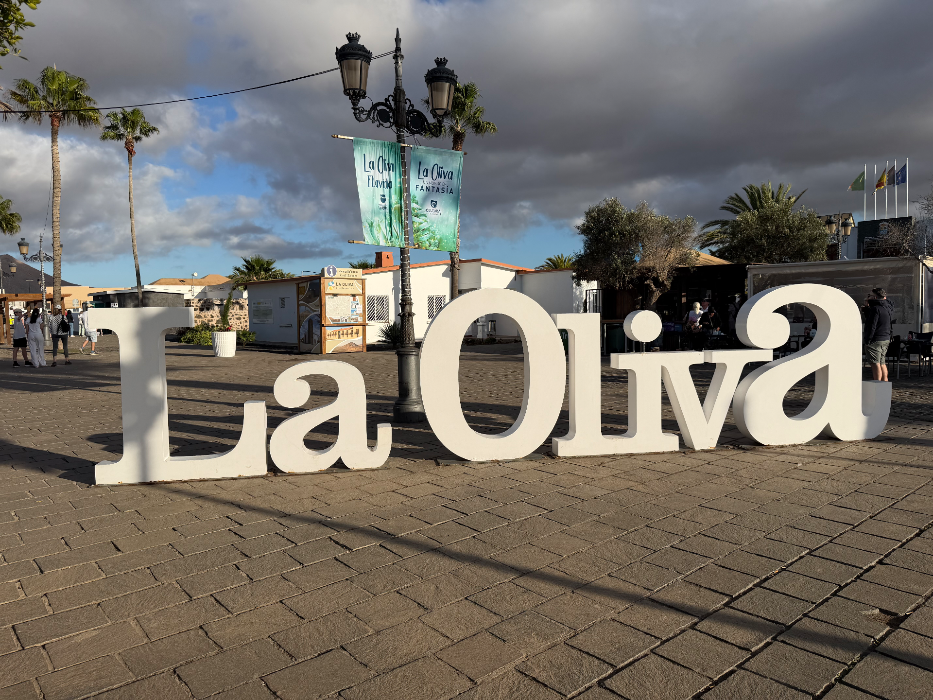 Large white 3D 'La Oliva' sign on a cobblestone plaza in Fuerteventura, with palm trees, a vintage lamppost, and dramatic clo